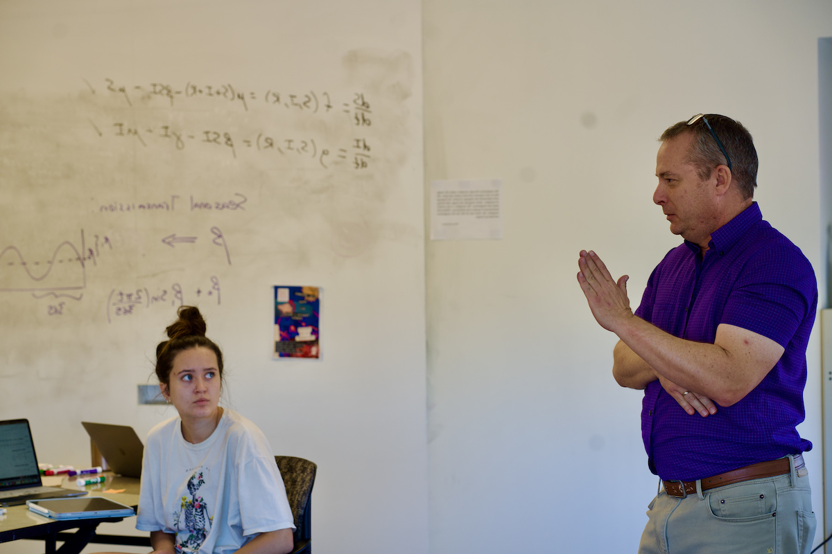 A male professor in a blue dress shirt explaining a concept of a class
